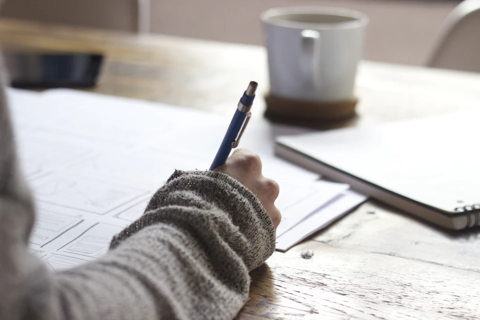Hands writing notes beside a white cup on a wooden table. Conceptual stock photograph.
